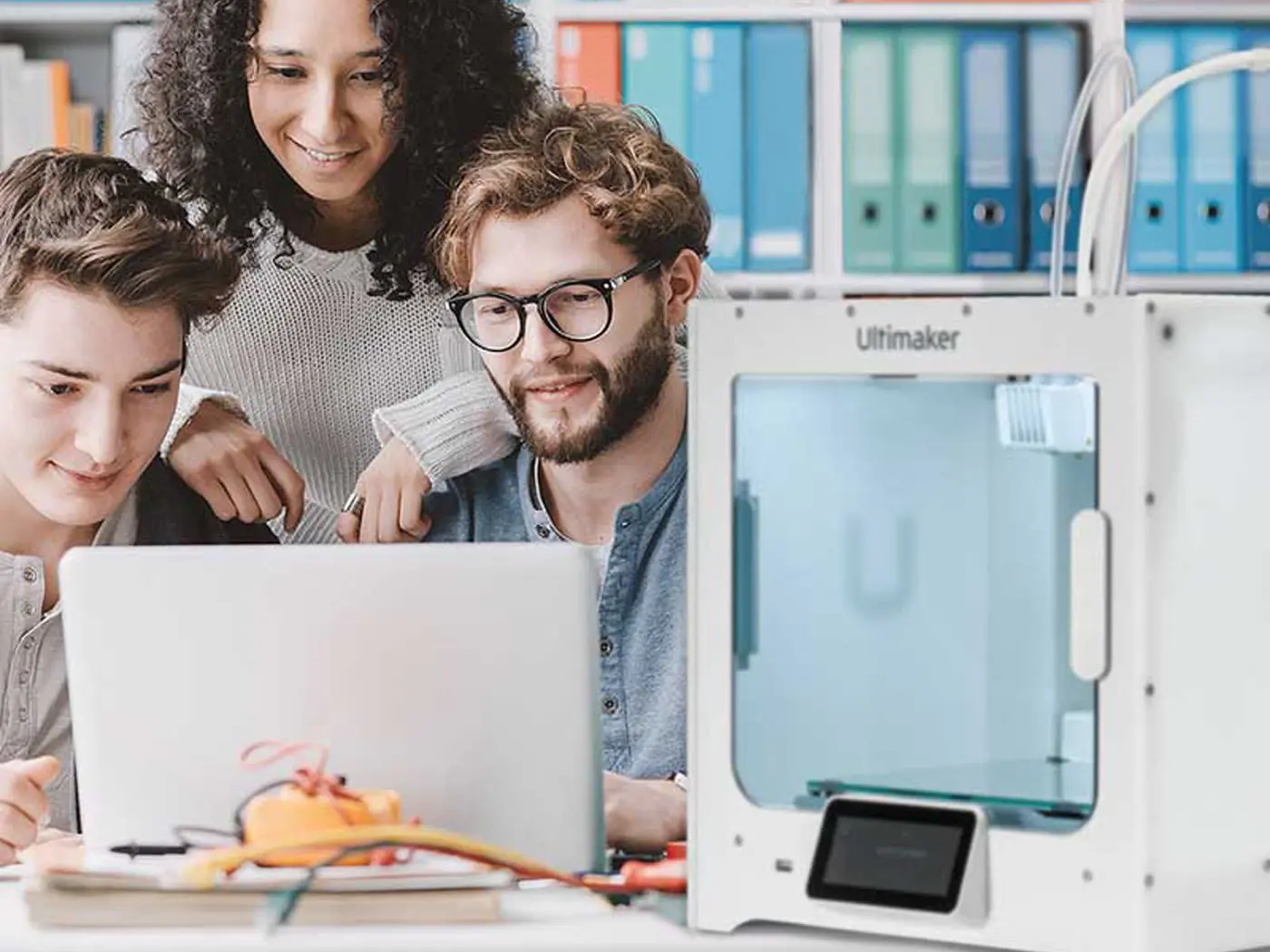 Drei Studierende schauen auf einem Laptop der mit einem 3D Drucker verbunden ist